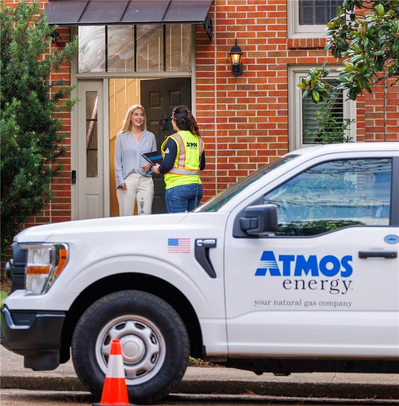 Atmos Energy employee wearing a safety vest speaks with a smiling homeowner at the front door of a brick house. An Atmos Energy truck is parked in front with a safety cone nearby.